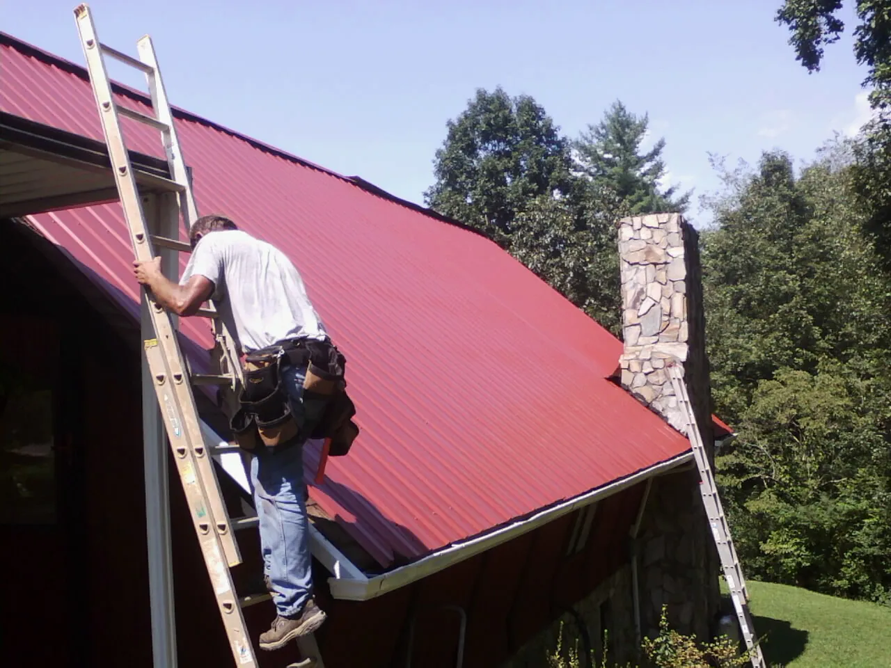 Roofer on a ladder beside a red metal roof and stone chimney — Soffit & Fascia Repair by Roofing Brothers in Frankton